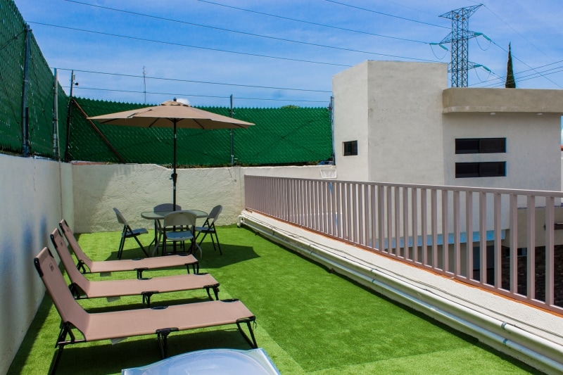 An open solarium features several lounge chairs arranged on artificial grass, accompanied by a round table and umbrella for shade. The area is surrounded by low walls, providing a private outdoor space with clear blue skies above.