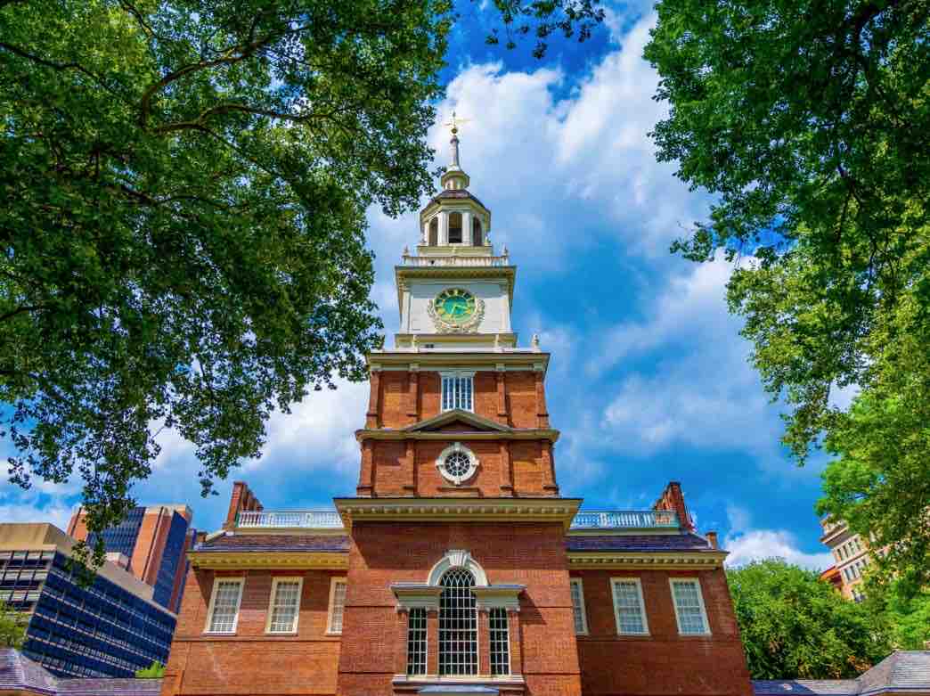 A historic brick building stands prominently against a backdrop of bright blue skies and fluffy white clouds. Lush green trees frame the scene, highlighting the tall clock tower topped with a gold weathervane. The structure features multiple windows and distinctive architectural details.