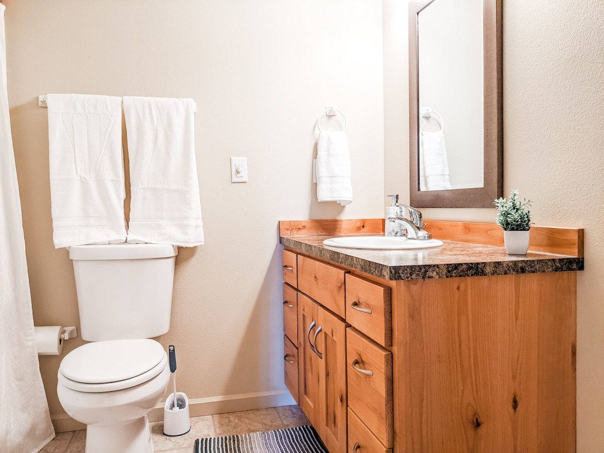 A clean bathroom is presented, featuring a wooden vanity with a granite countertop and a mirror above. The toilet is neatly positioned beside the vanity. Two white towels hang on nearby hooks, contributing to the overall simplicity and functionality of the space.