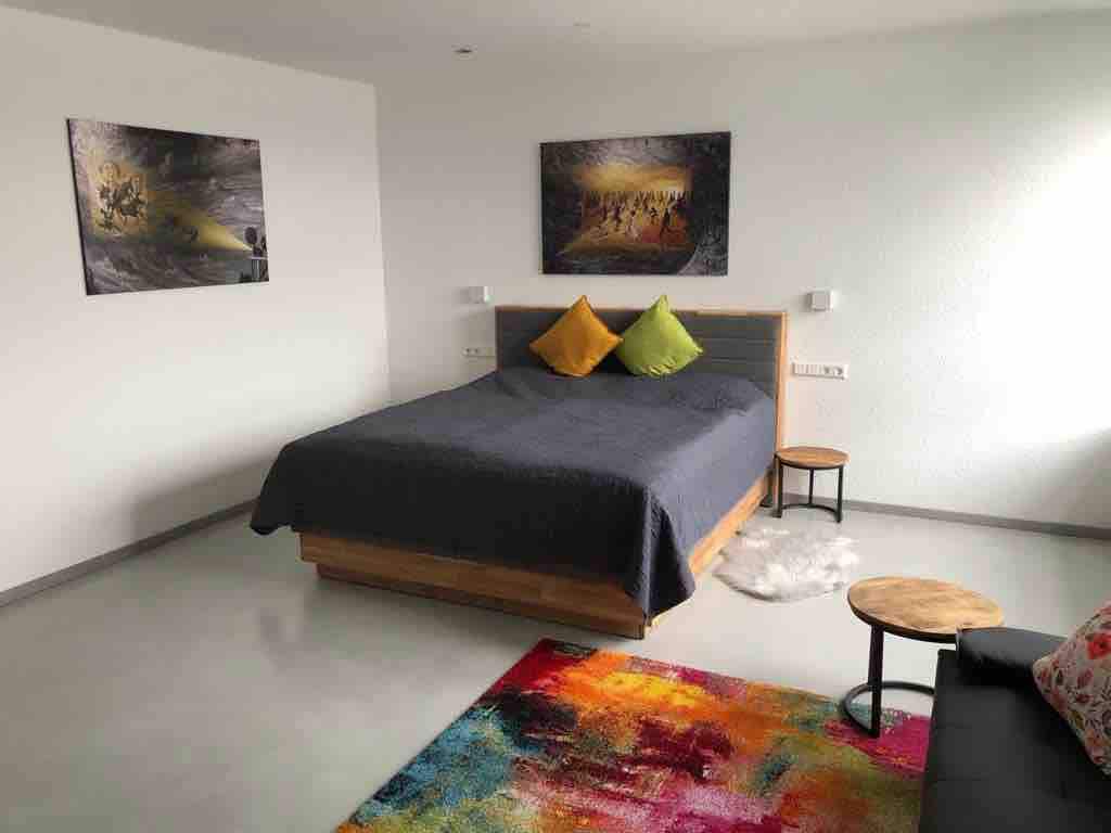 A spacious room is furnished with a comfortable bed featuring colorful cushions. Two wall-mounted lamps are positioned beside the bed, and two artworks adorn the white walls. A small side table complements the seating area, which includes a cozy sofa. A vibrant rug adds warmth to the floor.