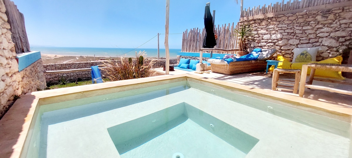 A private pool with a clear, sparkling surface reflects the sky, surrounded by a wooden deck. Colorful lounge chairs and a sun umbrella are positioned nearby, providing a comfortable area to relax while enjoying the expansive view of the ocean in the background.
