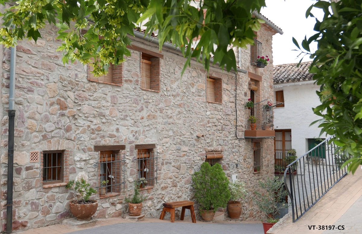 A rustic stone facade is presented, featuring several windows adorned with wooden shutters and potted plants. An inviting entrance path leads to the house, framed by greenery and traditional stone elements characteristic of the local architecture.