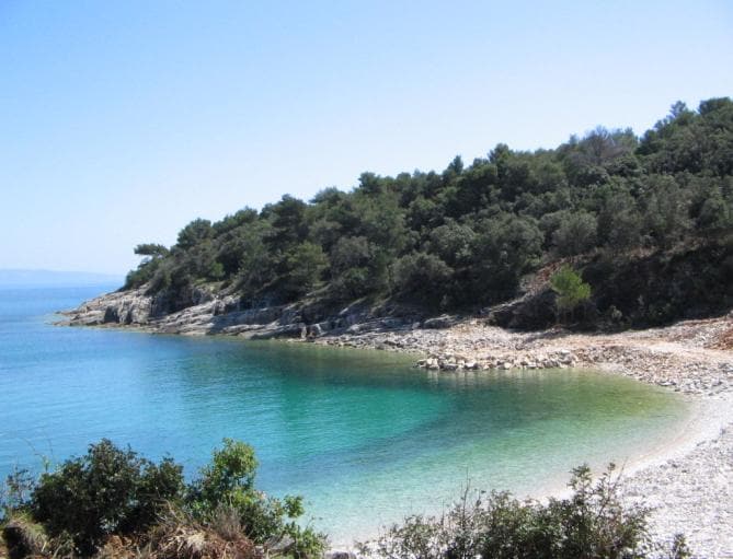 A serene beach is depicted with clear turquoise waters gently lapping against a pebbled shore. Lush greenery lines the coastline, providing a natural backdrop. The sky is bright and clear, enhancing the tranquil atmosphere of this coastal setting.
