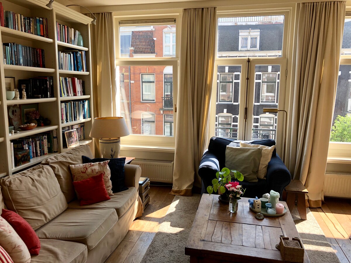 A bright living area is illuminated by natural light streaming through large windows. A comfortable sofa with colorful cushions is positioned beside a wooden coffee table, adorned with decorative items and a small plant. A bookshelf filled with books adds an inviting touch to the space.