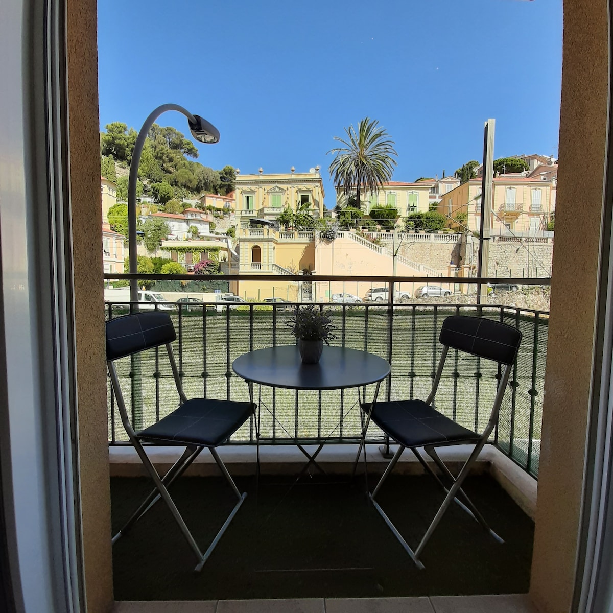 A small balcony is visible, featuring a round table and two folding chairs. A potted plant is centered on the table. In the background, a view of terraced houses and palm trees against a clear blue sky is highlighted.
