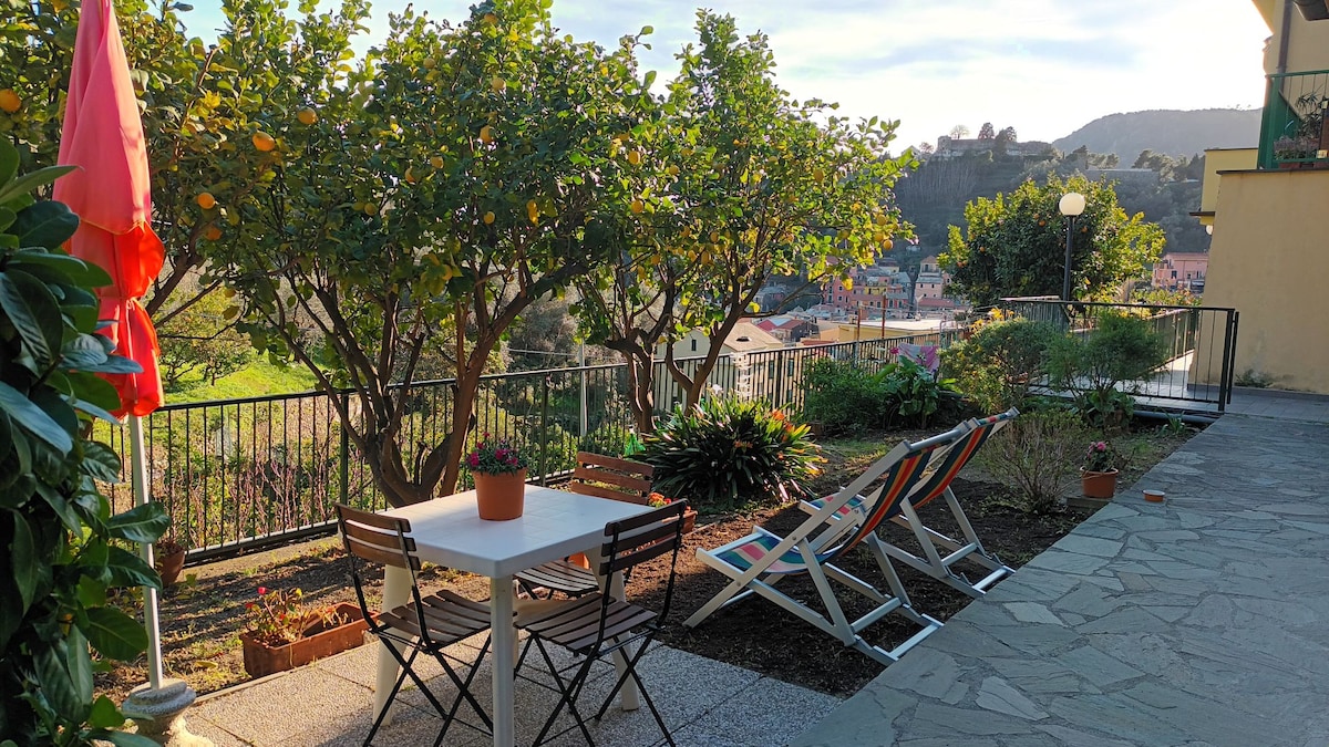 A garden space is presented with a small table and four chairs, accompanied by lounge chairs positioned on a gravel surface. Lush greenery surrounds the area, with lemon trees offering shade. Scenic views of the nearby landscape are visible in the background.