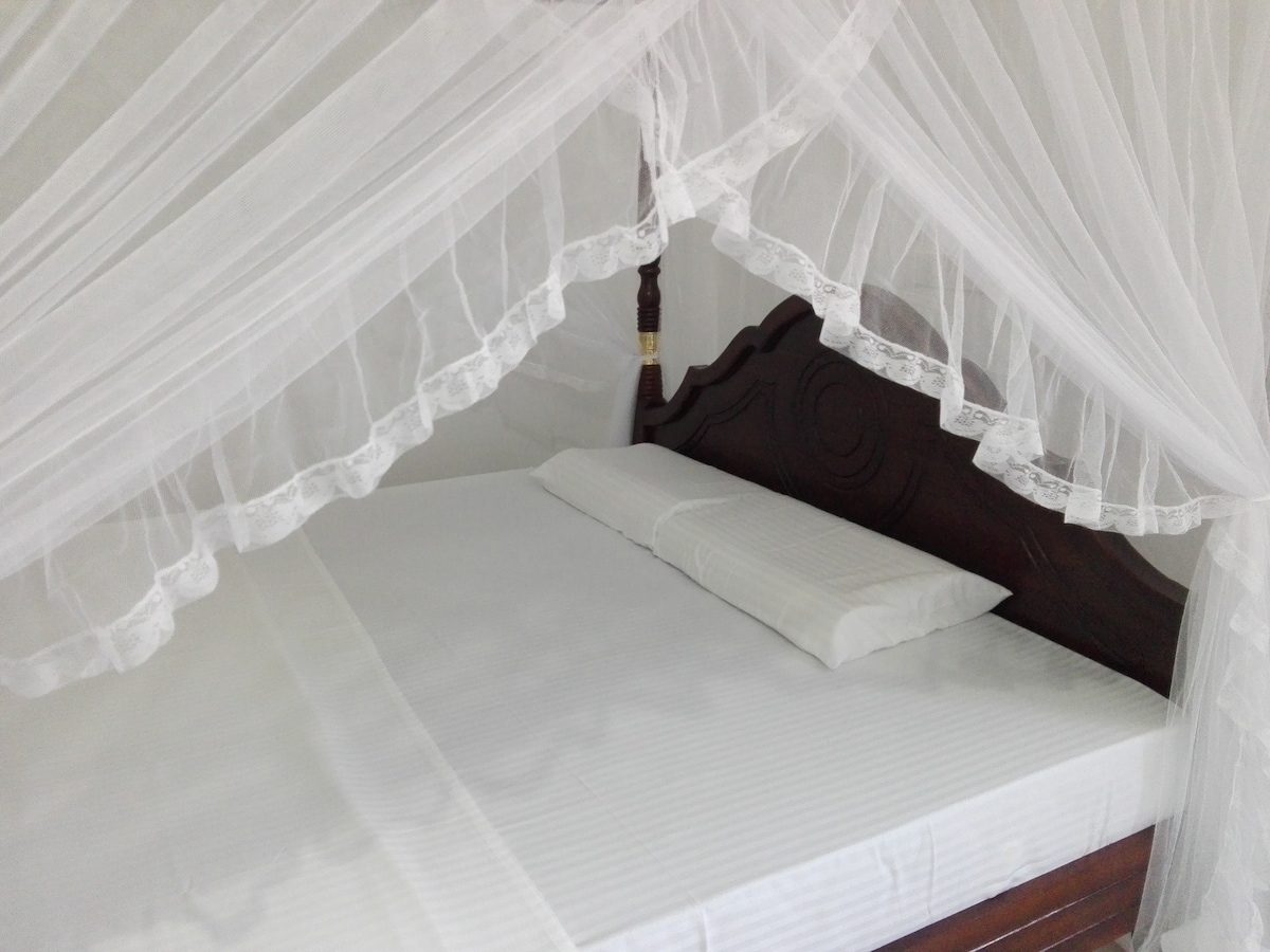 A spacious bed is dressed in crisp white linens, draped with a sheer canopy that features delicate lace trim. A pair of pillows rests at the headboard, inviting a restful night. Soft, diffused light enhances the serene atmosphere of the sleeping area.