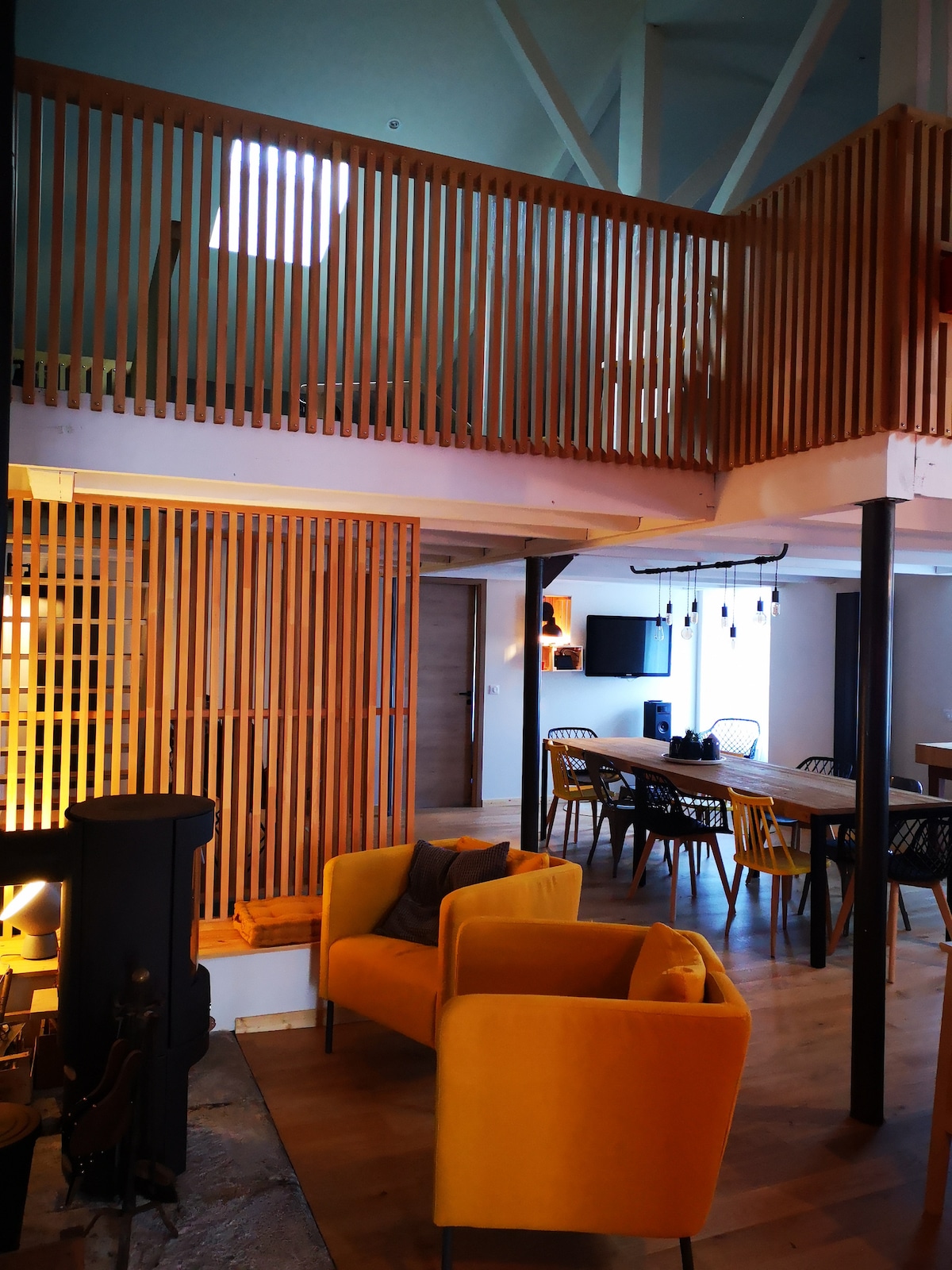 A spacious living area is showcased with a warm wooden staircase leading to the mezzanine. Yellow armchairs provide comfortable seating near a modern wood stove, while a dining table is visible in the background, accompanied by stylish black chairs. Natural light filters through a skylight.