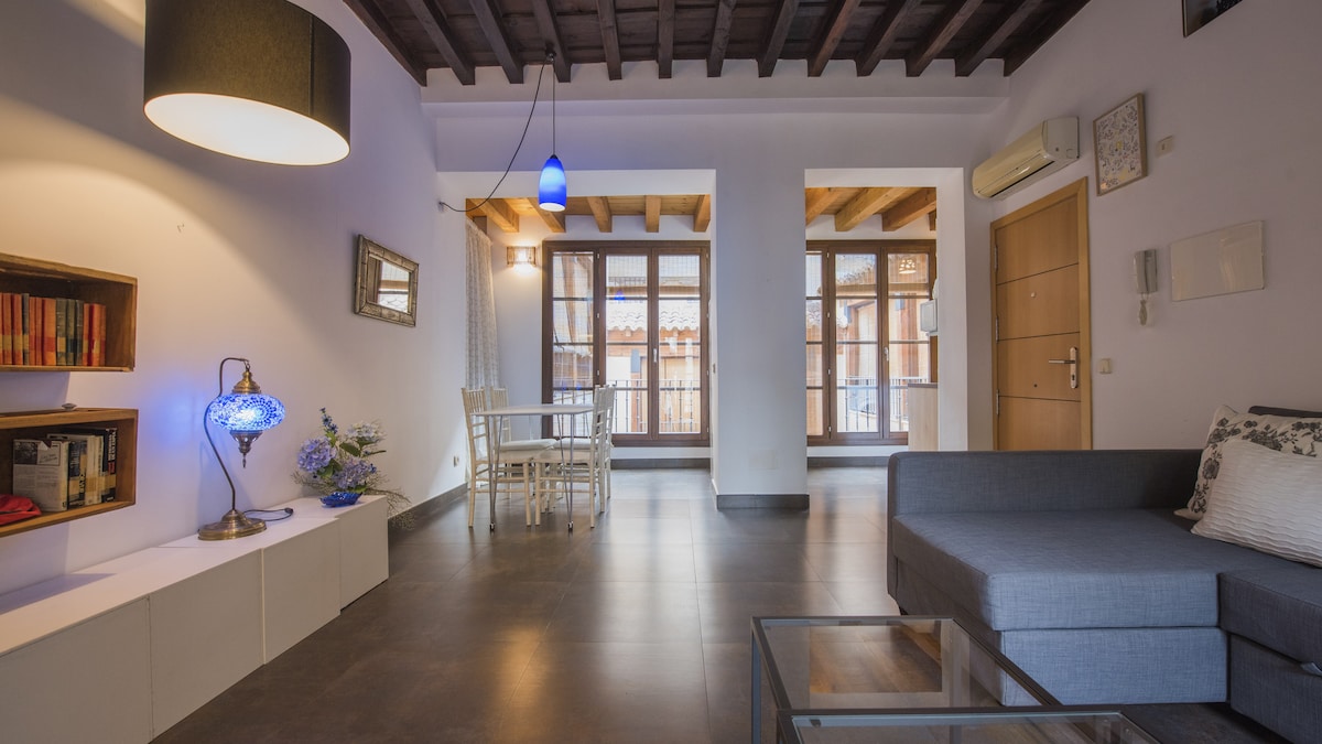 An open living area is presented with exposed wooden beams and natural light streaming through large windows. A comfortable sofa occupies one side, while a dining table and chairs are positioned near the window. Shelving displays books and decorative items, enhancing the room's charm.
