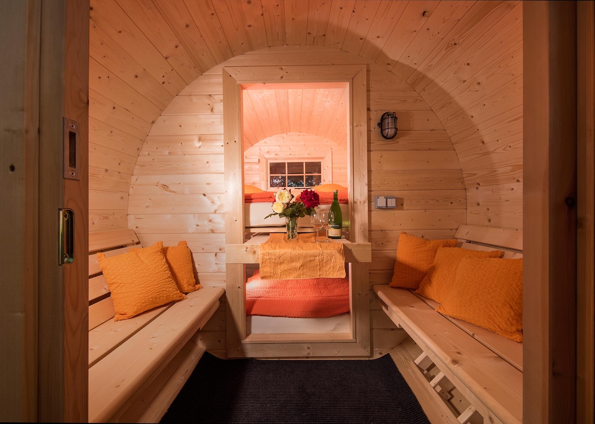 The interior of the barrel-shaped accommodation features wooden walls and a warm ambiance. Two benches are positioned on either side, with decorative orange cushions. A table is set at the center, accompanied by a vase of flowers, with a glimpse of the sleeping area visible in the background.