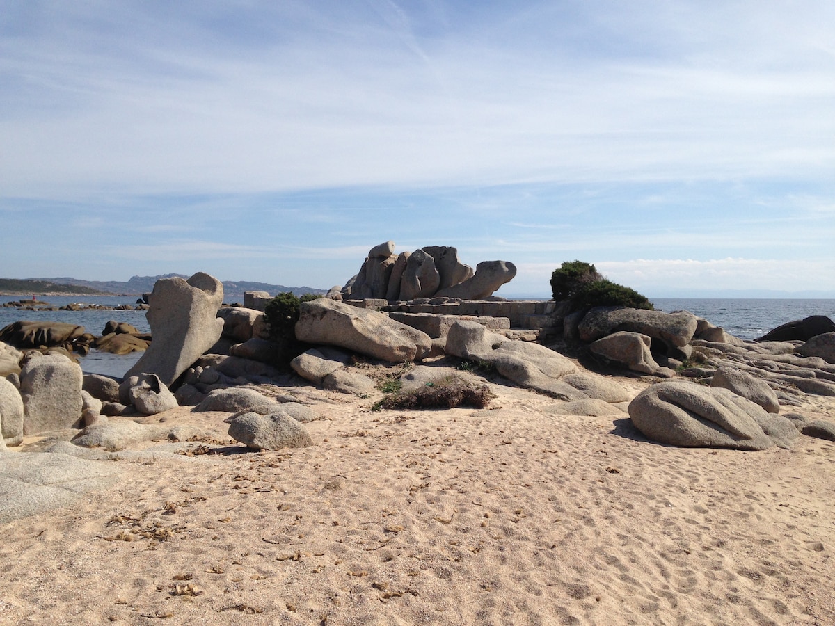 A sandy beach is visible, extending towards rocky formations by the water's edge. Smooth, rounded stones are scattered across the beach, while the calm sea reflects the sky. In the background, distant hills add to the serene coastal landscape.