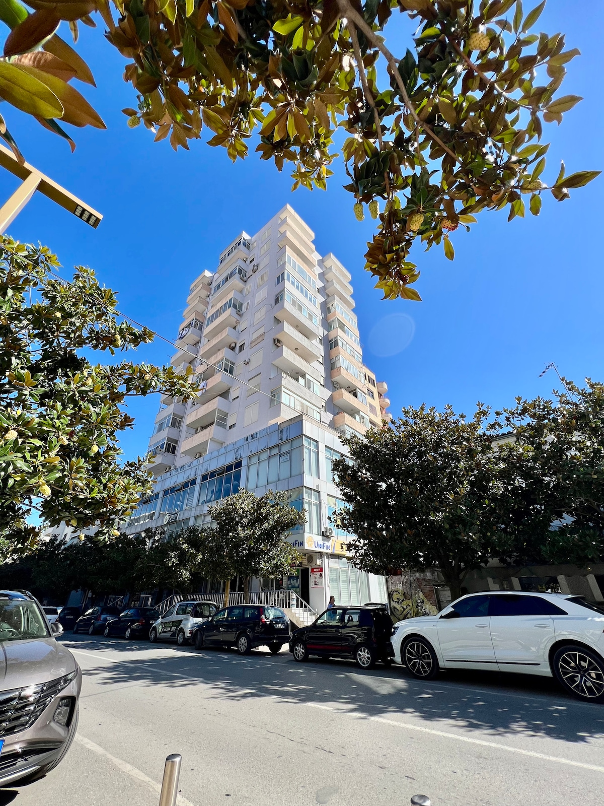 A modern multi-story building is shown from street level, with several floors featuring glass balconies and large windows. The structure is surrounded by leafy trees that provide shade to parked cars along the street, creating a blend of urban and natural elements.