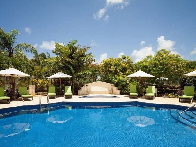 A communal swimming pool is surrounded by lush greenery and well-maintained gardens. Comfortable loungers with bright green cushions are placed under large umbrellas, offering shaded relaxation areas. Clear blue skies and a few wispy clouds enhance the bright outdoor setting.