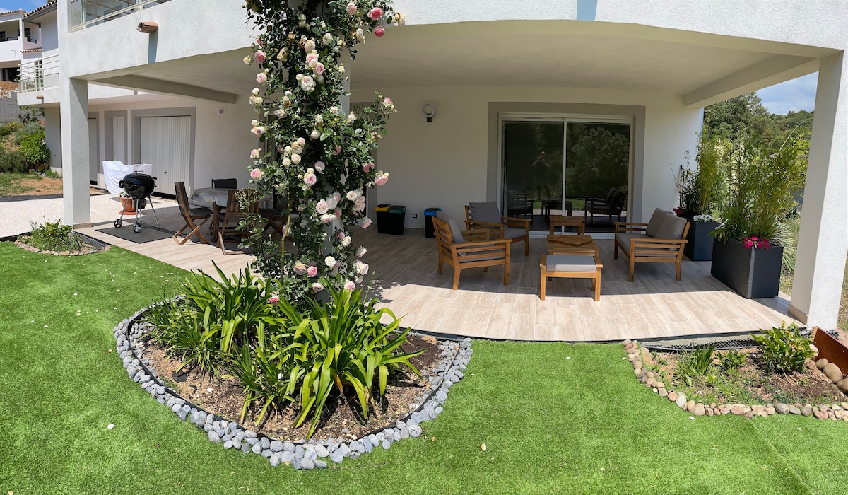 A spacious covered terrace features a wooden seating area and a dining set, surrounded by well-maintained greenery. A barbecue grill is positioned nearby. The terrace provides an inviting outdoor space, enhanced by flowering plants and decorative stone borders.