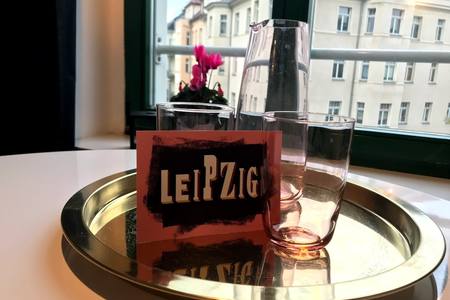 A silver tray displays two transparent glasses and a matching pitcher, accompanied by a card that reads 'LEIPZIG' in bold lettering. A view of a blooming pink plant is seen through a large window, framed by green accents.