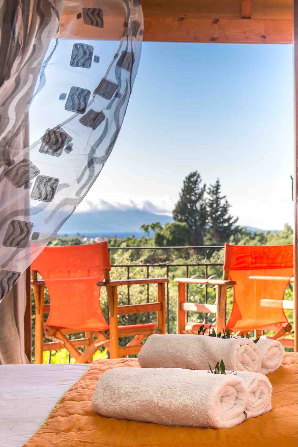 A balcony view features comfortable orange chairs overlooking a serene landscape. Rolled towels are placed neatly on a soft surface, complementing the space. A sheer curtain gently frames the scene, allowing natural light to brighten the area.