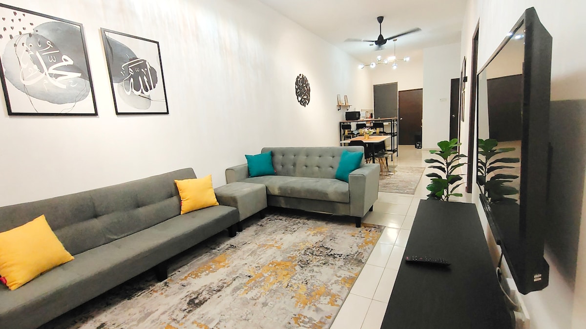 The living area features a modern design with two gray sofas adorned with yellow cushions. A large area rug lies underfoot, complementing the neutral walls. A wall-mounted television is positioned across from the seating, and art pieces are displayed above the sofas, adding character to the space.