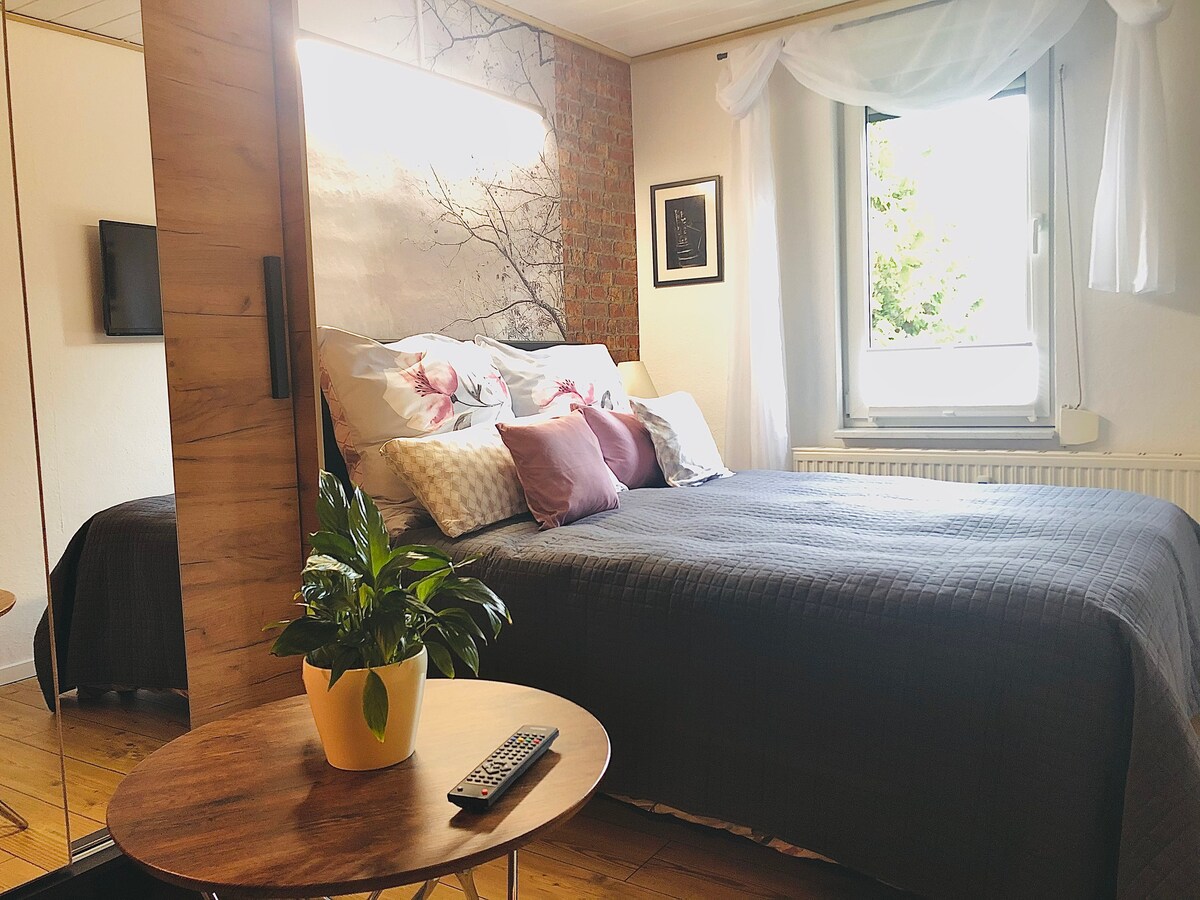 A sleeping area is presented with a spacious bed adorned with decorative pillows and a textured blanket. Natural light enters through a window, creating a soft glow. A small table with a plant and remote control is positioned nearby, adding to the inviting setup.