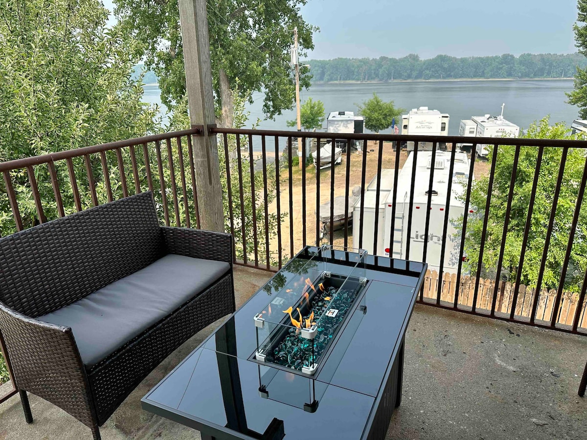 A balcony features a dark wicker seating area with a comfortable chair and a glass-topped table. The view overlooks the serene Mississippi River, with trees lining the banks and a glimpse of nearby recreational vehicles.