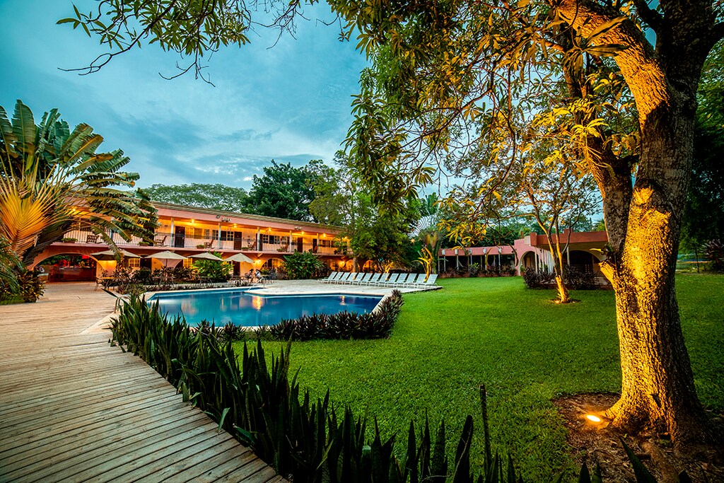 A serene view of the swimming pool surrounded by lush tropical gardens. Lounge chairs are positioned by the water, and a wooden pathway leads through the greenery. The colonial-style building is partially visible in the background, set against a calming sky.