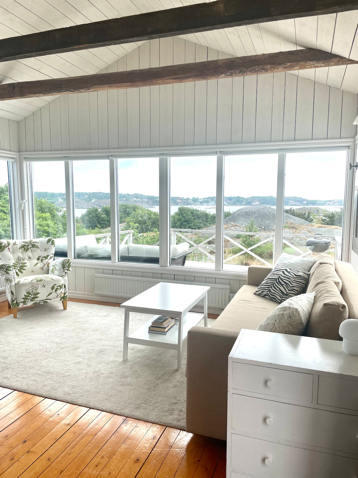 The living room features large windows offering a view of the sea and surrounding landscape. A light-colored sofa and a white coffee table are placed on a soft rug. A patterned armchair adds a touch of elegance to the space, creating a bright atmosphere.