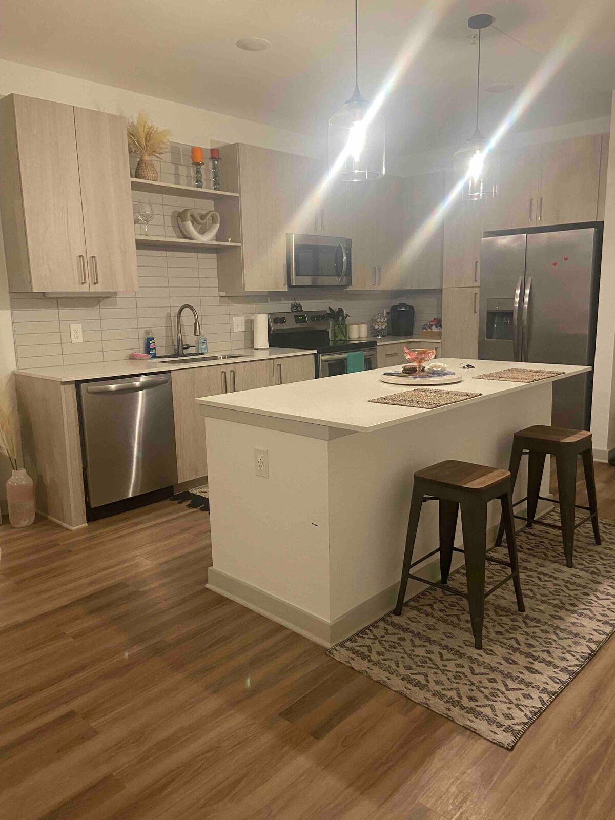 A modern kitchen is featured, equipped with stainless steel appliances and a large island with seating for two. Elegant overhead lighting illuminates the space, which includes light wood cabinetry and a tiled backsplash. A decorative rug lies beneath the island, adding a cozy touch.