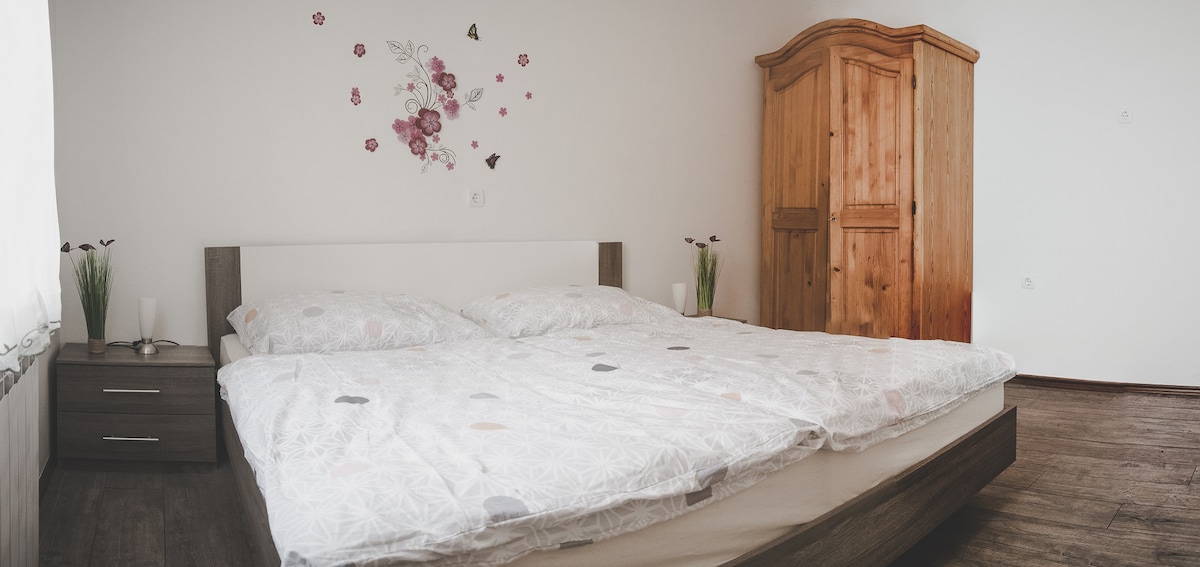 A cozy bedroom features a spacious bed with soft bedding and light floral accents. A wooden wardrobe stands against the wall, complemented by a nightstand on either side, each adorned with decorative plants. Natural light gently illuminates the room through a nearby window.