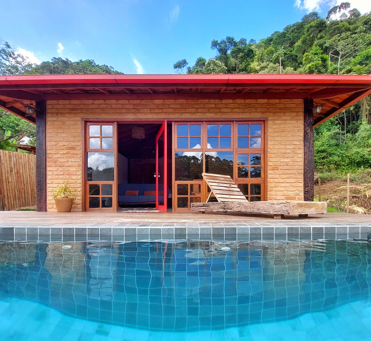 A charming exterior of a rustic building features large glass doors opening to a patio. A cozy wooden lounge chair sits near the edge of a tranquil infinity pool, surrounded by lush greenery and mountains in the background.