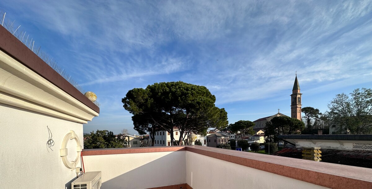 A terrace view displays a tranquil scene featuring a tall tree and a historic church steeple against a clear blue sky. The surrounding rooftops add to the sense of openness and connection to the historic ambience of the area.