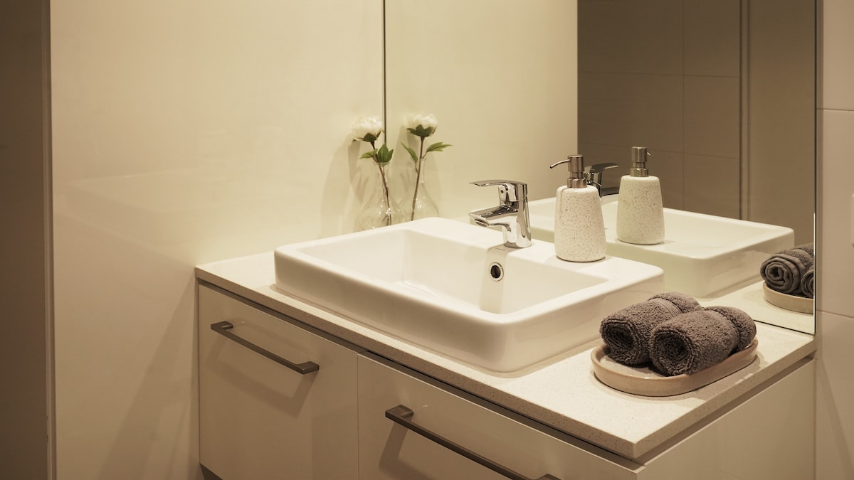 A modern bathroom sink area is presented with a sleek countertop, featuring a rectangular basin and contemporary faucet. Neatly rolled gray towels are arranged on a tray, alongside minimalist accessories. Reflections in the mirror add depth to the space, enhanced by soft lighting and subtle floral accents.