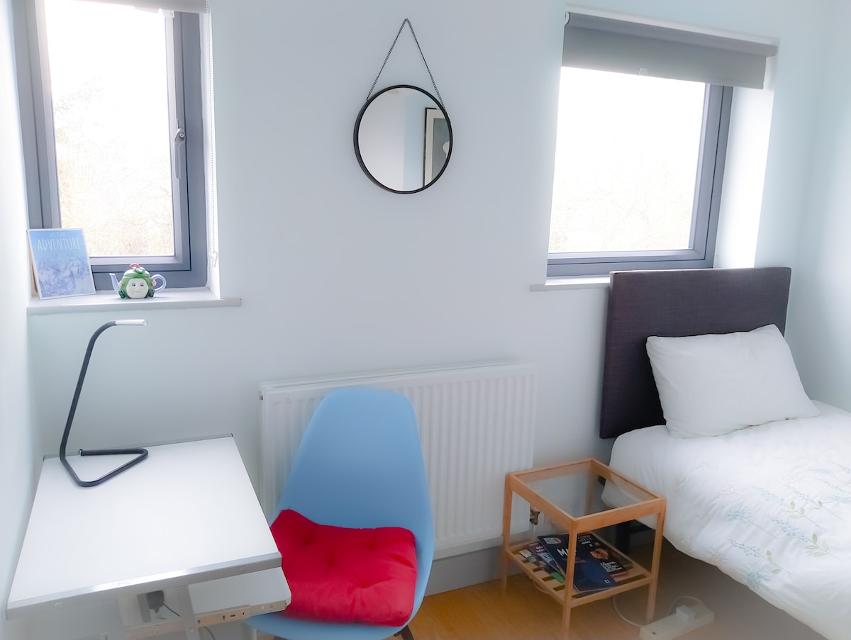 A newly decorated bedroom is presented, featuring a single bed with crisp bedding. A small bedside table is positioned next to the bed, while a desk and colorful chair occupy one corner. Two aluminium windows allow natural light to fill the space, complemented by a round mirror on the wall.
