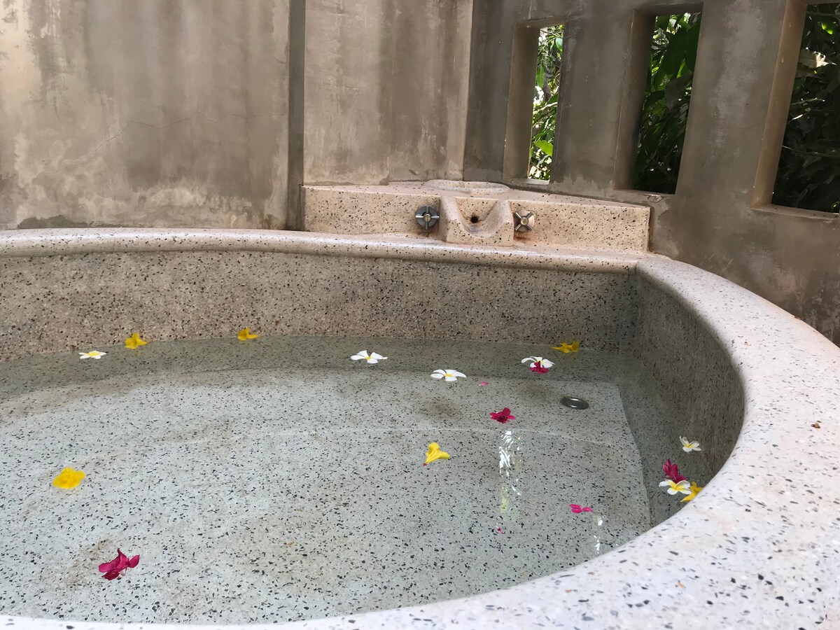 A circular soaking tub with smooth, speckled stone surrounds is filled with clear water. Colorful flower petals float gently on the surface, adding a touch of natural beauty. Light filters in through nearby openings, creating a calm and serene atmosphere.