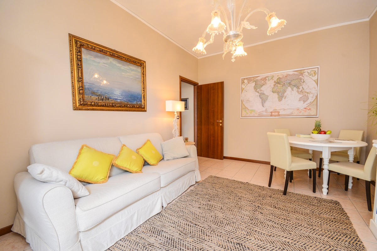 A light and inviting living area features a white sofa adorned with yellow cushions, accompanied by a round dining table surrounded by four chairs. A decorative map is displayed on the wall, and a chandelier adds brightness to the space.