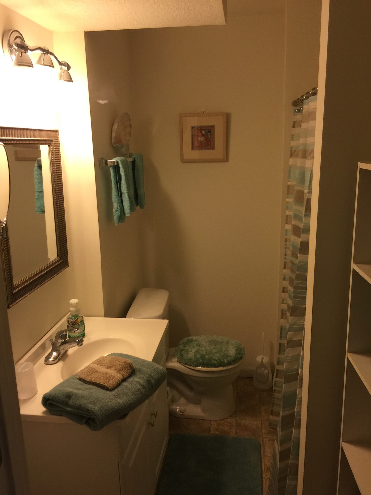 A compact bathroom features light-colored walls and tiled flooring. A mirror is mounted above a white vanity with a towel and soap dispenser placed nearby. A shower with a patterned curtain is visible, along with neatly arranged towels in soft tones.