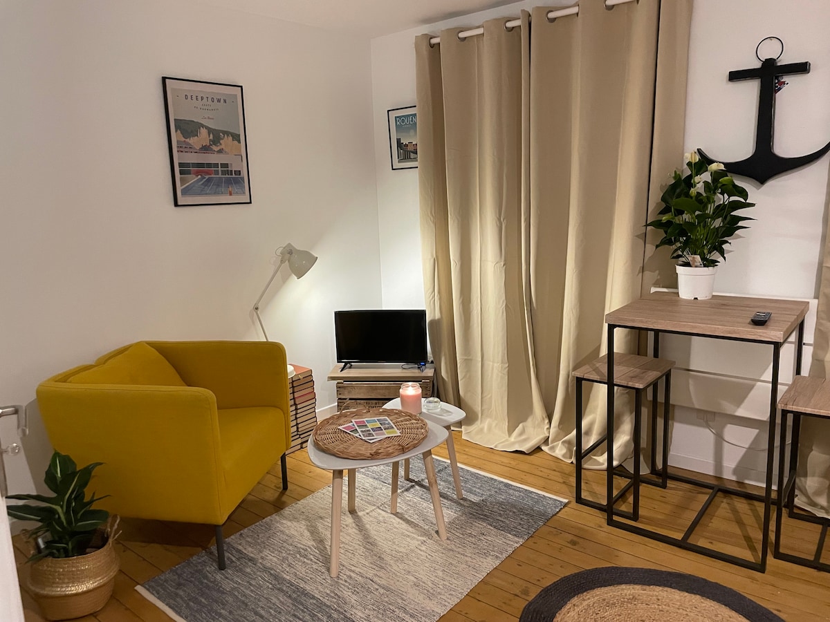 A cozy living area features a vibrant yellow armchair and a small round wooden table. A compact television sits on a stand next to a decorative plant. Light-colored curtains frame a window, allowing natural light to enhance the warm ambiance of the space.