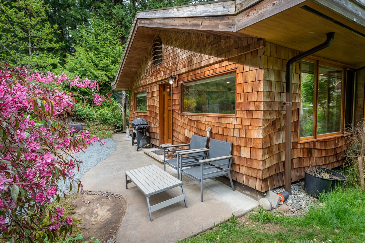 An inviting entrance to the Nautilus Suite is framed by lush greenery and vibrant pink flowers. A patio area features outdoor seating with two chairs and a low table, adjacent to a barbecue. Large windows allow natural light to highlight the wooden shingles of the exterior.