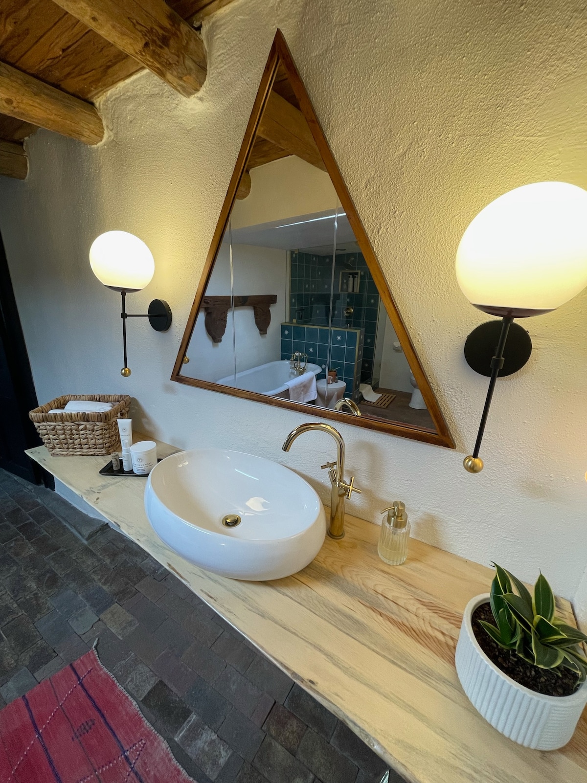 A modern bathroom vanity features a sleek, white basin resting on a natural wood countertop. A triangular mirror reflects the space, illuminated by two spherical wall sconces. A woven basket and potted plant add subtle elements of warmth and greenery.