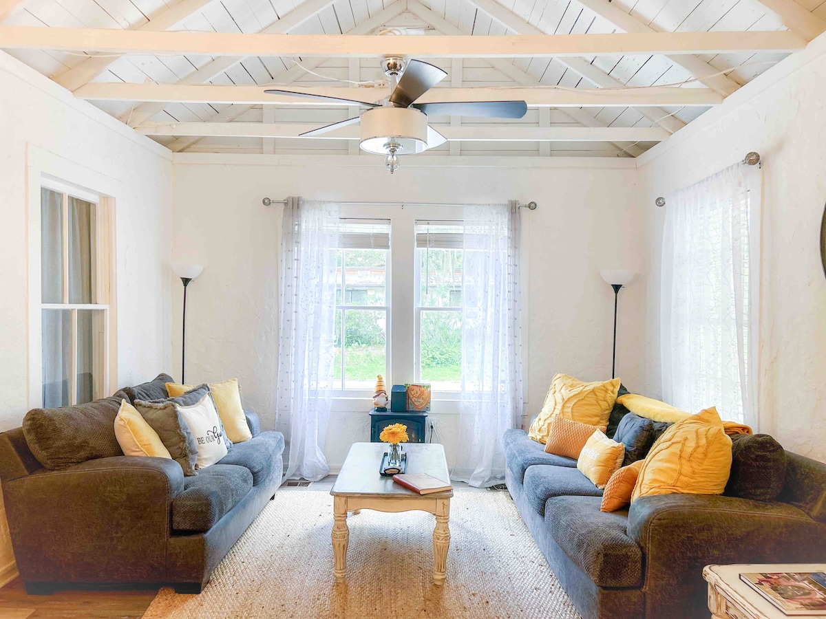 A welcoming living room is highlighted by two comfortable gray sofas adorned with yellow cushions. Natural light floods the space through large windows framed by sheer curtains, while a white coffee table lies in the center, complemented by decorative elements. The ceiling features exposed beams, enhancing the room's charm.
