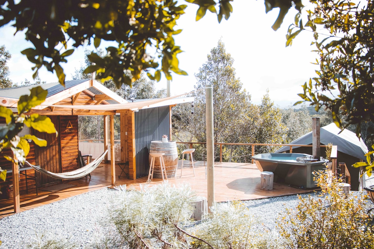 An outdoor area is presented, featuring a wooden deck surrounded by greenery. A hammock hangs between wooden posts, and a stylish hot tub is positioned nearby. An outdoor kitchen is visible, complemented by a small seating area, all designed for a nature-friendly experience.