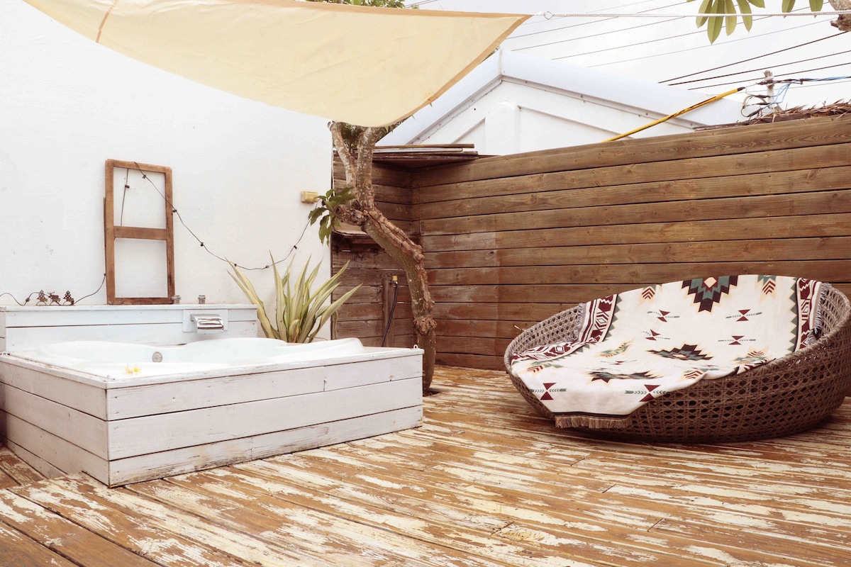 An outdoor area features a wooden deck with a spacious circular lounge chair, adorned with a patterned throw. A white hot tub is positioned nearby, complemented by potted greenery and ambient lighting, creating an inviting atmosphere for relaxation.