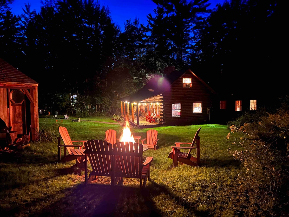 A spacious outdoor area is illuminated by a firepit surrounded by quality Adirondack chairs. The warm glow of the fire contrasts with the deep blue night sky, while the inviting cabin features multiple windows that offer glimpses of cozy indoor seating.