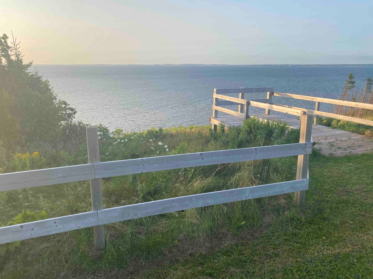A wooden fence outlines a grassy area leading to a scenic view of Orwell Bay, where soft blue waters meet the horizon. Sunlight gently illuminates the space, highlighting wildflowers and vegetation along the pathway. The serene landscape evokes a sense of tranquility.