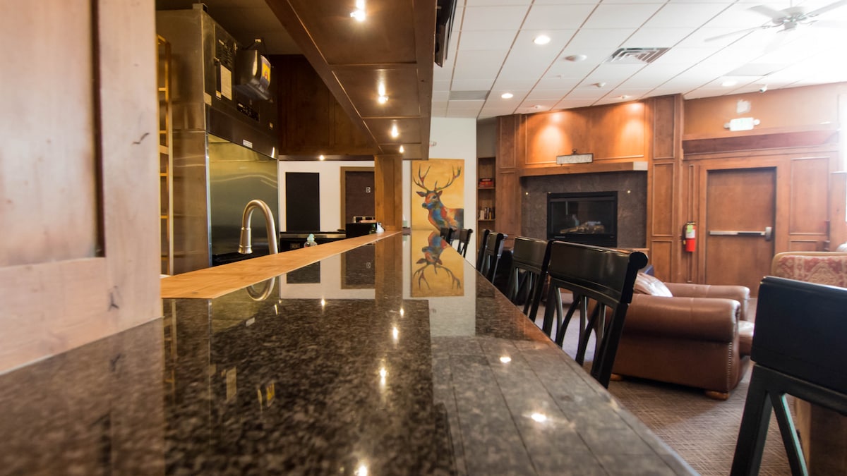 A well-appointed common area features a polished granite countertop with a bar sink. The space includes comfortable seating, a cozy fireplace area, and modern kitchen appliances visible in the background. Soft lighting enhances the welcoming atmosphere of the room.