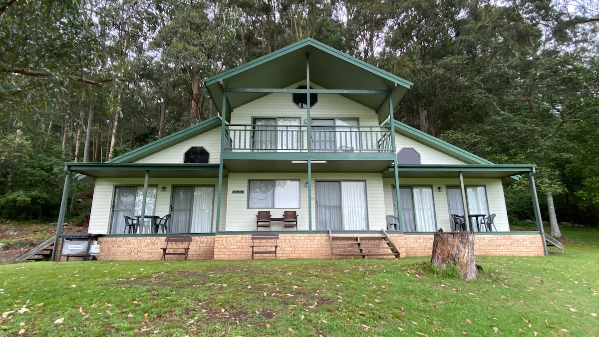 The two-story villa features a green gabled roof and light-colored siding, surrounded by natural greenery. A spacious front porch offers seating with wooden chairs and tables. Large glass doors provide access to the interior spaces, enhancing the connection to the outdoors.