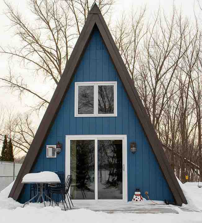 A striking A-frame cabin is depicted, featuring a vibrant blue exterior complemented by dark trim. Large windows invite natural light, while a small patio area with two chairs is visible. Snow blankets the surrounding ground, enhancing the winter landscape.