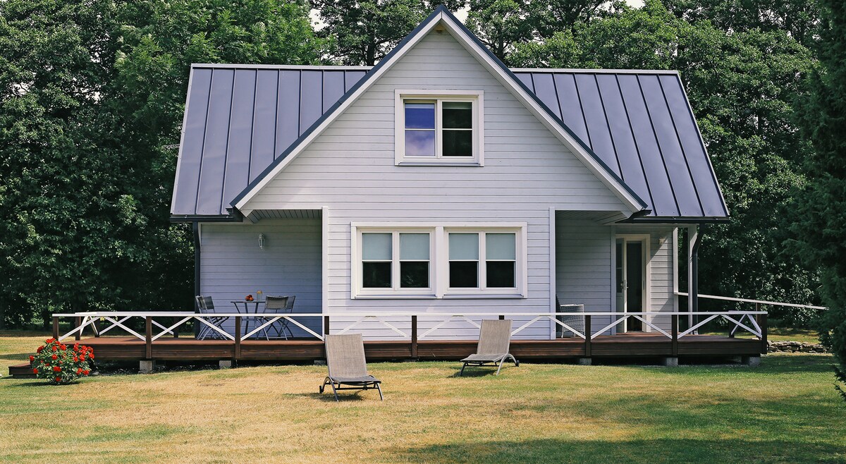 A spacious holiday home is set amidst lush greenery, featuring a light-colored facade and a distinctive sloped roof. A wooden deck with lounge chairs provides an inviting outdoor space, while large windows allow natural light to fill the interior.