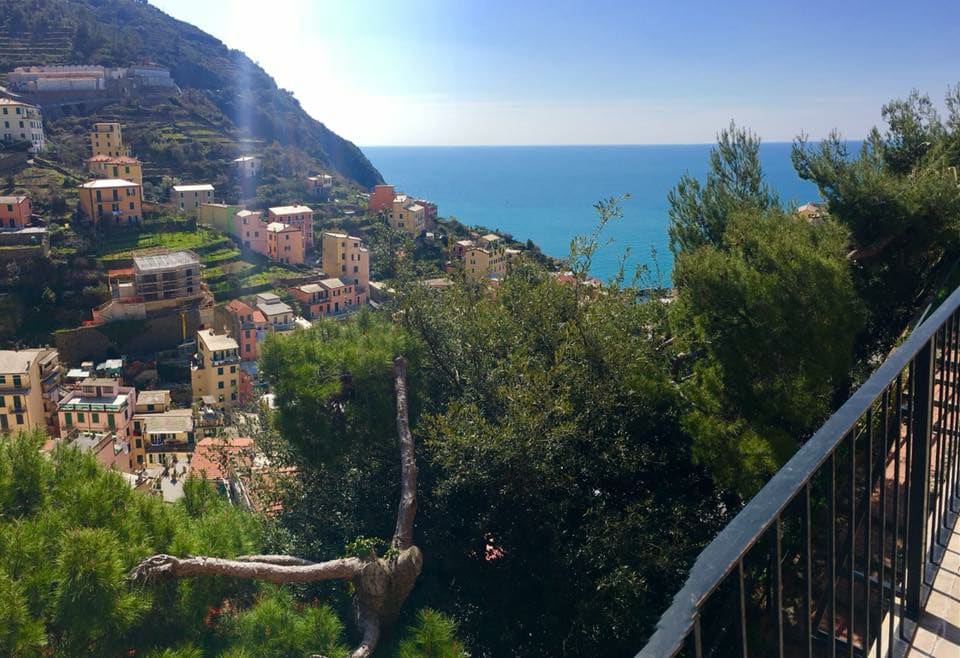 A panoramic view of a coastal village is presented, featuring colorful buildings nestled among lush greenery. The shimmering blue sea stretches out towards the horizon, and the surrounding hills provide a scenic backdrop to the tranquil landscape.
