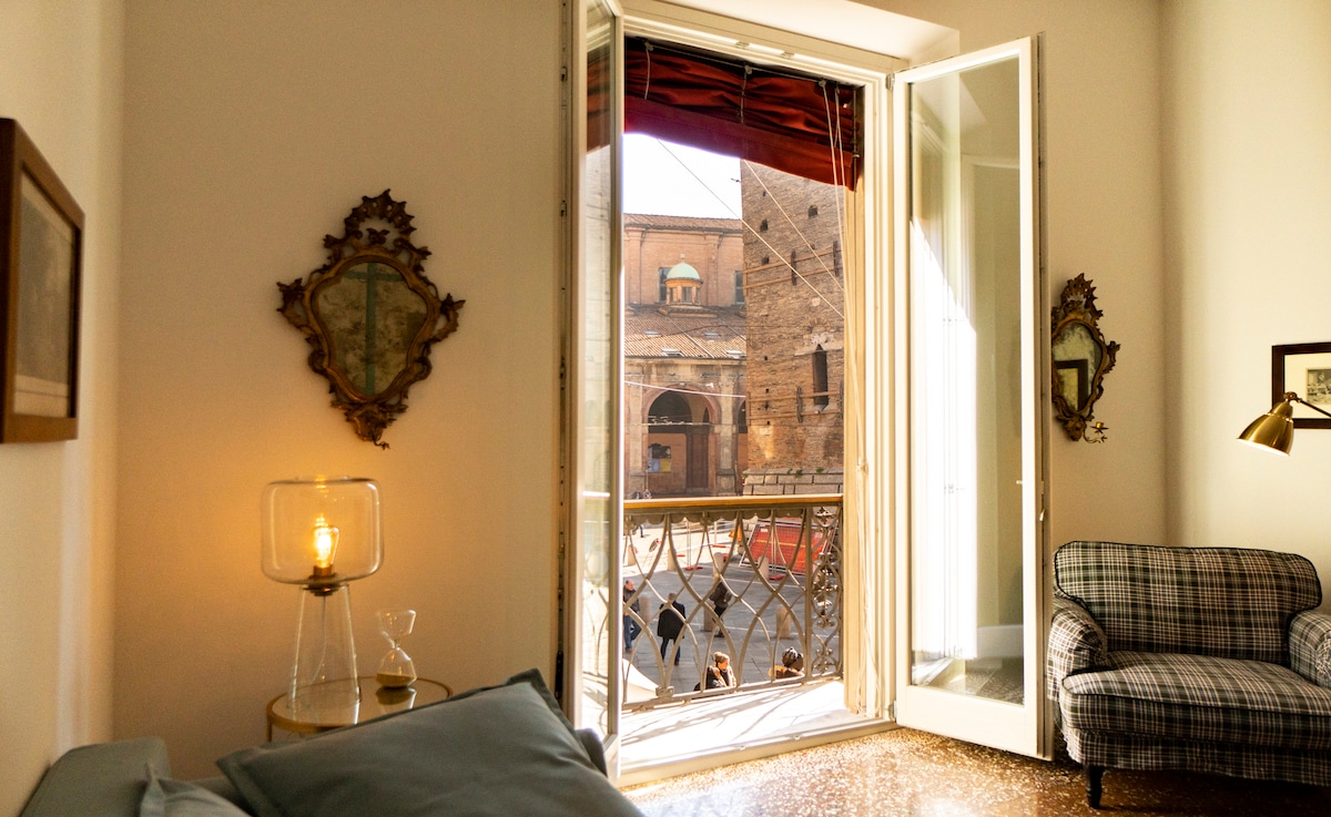 A well-lit room features large open double doors leading to a balcony. Traditional decor includes framed artworks on the walls and a cozy checkered sofa. Natural light floods the space, highlighting the polished floor and creating an inviting environment.