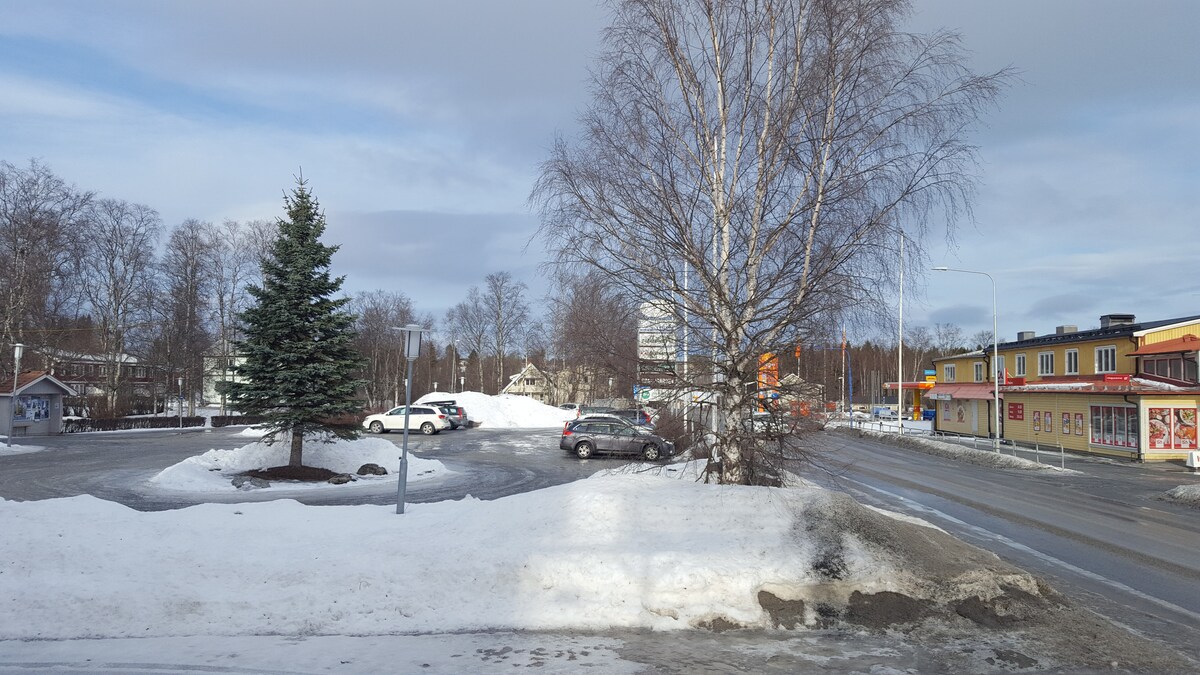 A winter scene features a snow-covered parking area surrounded by trees. A green evergreen tree stands on the left, while a few vehicles are parked nearby. The road in the foreground is partially cleared of snow, leading to colorful buildings on the right.