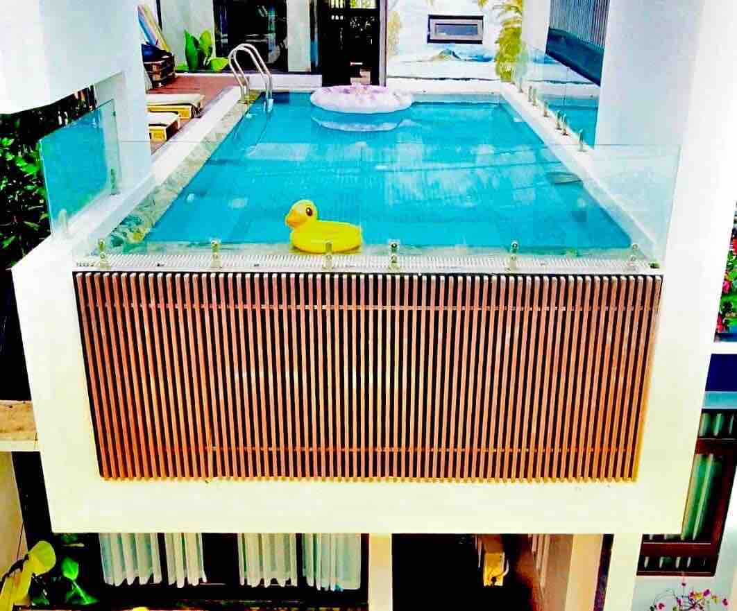 An elevated rooftop pool is surrounded by glass railings, offering a clear view of the water. A yellow floatie and lush greenery enhance the serene environment. The pool's surface reflects light, adding to the tranquil setting.